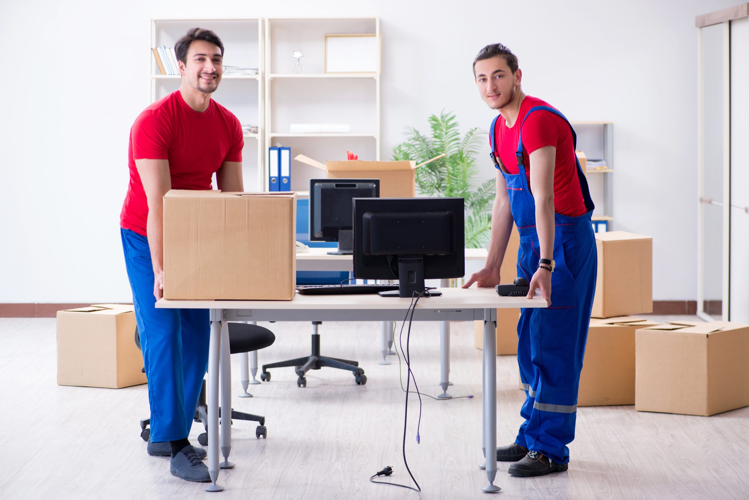 Two men in blue and red movers’ outfits place a desk with a computer in a new office.